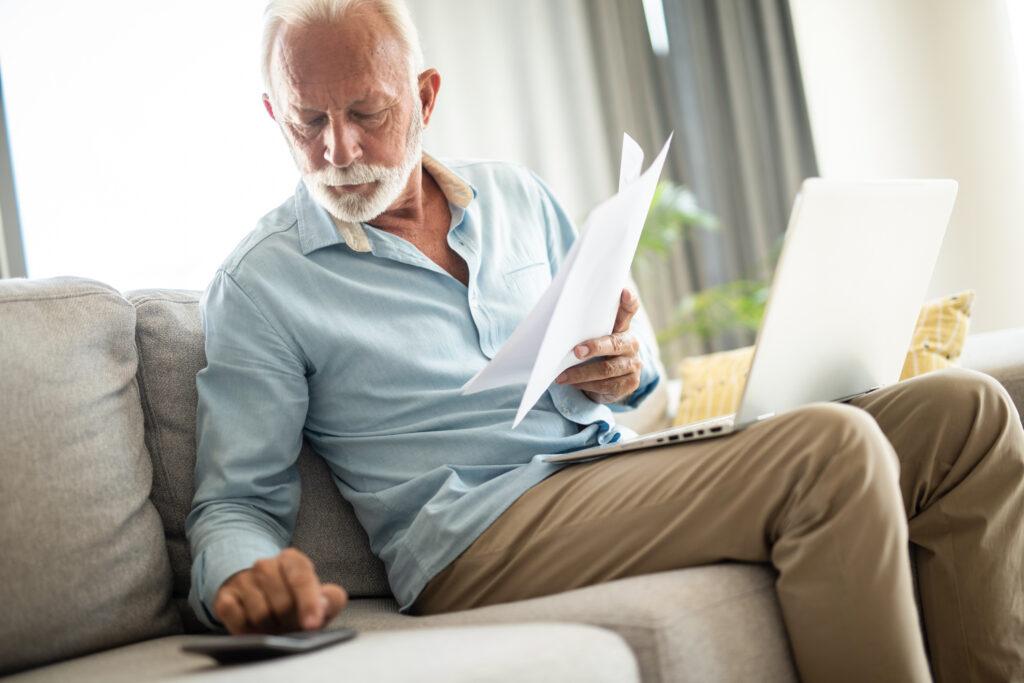 senior-man-couch-docs-calc-laptop-gettyimages-1343962734